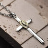 A silver cross amulet necklace with a gold-colored pendant resting on a light brown surface.