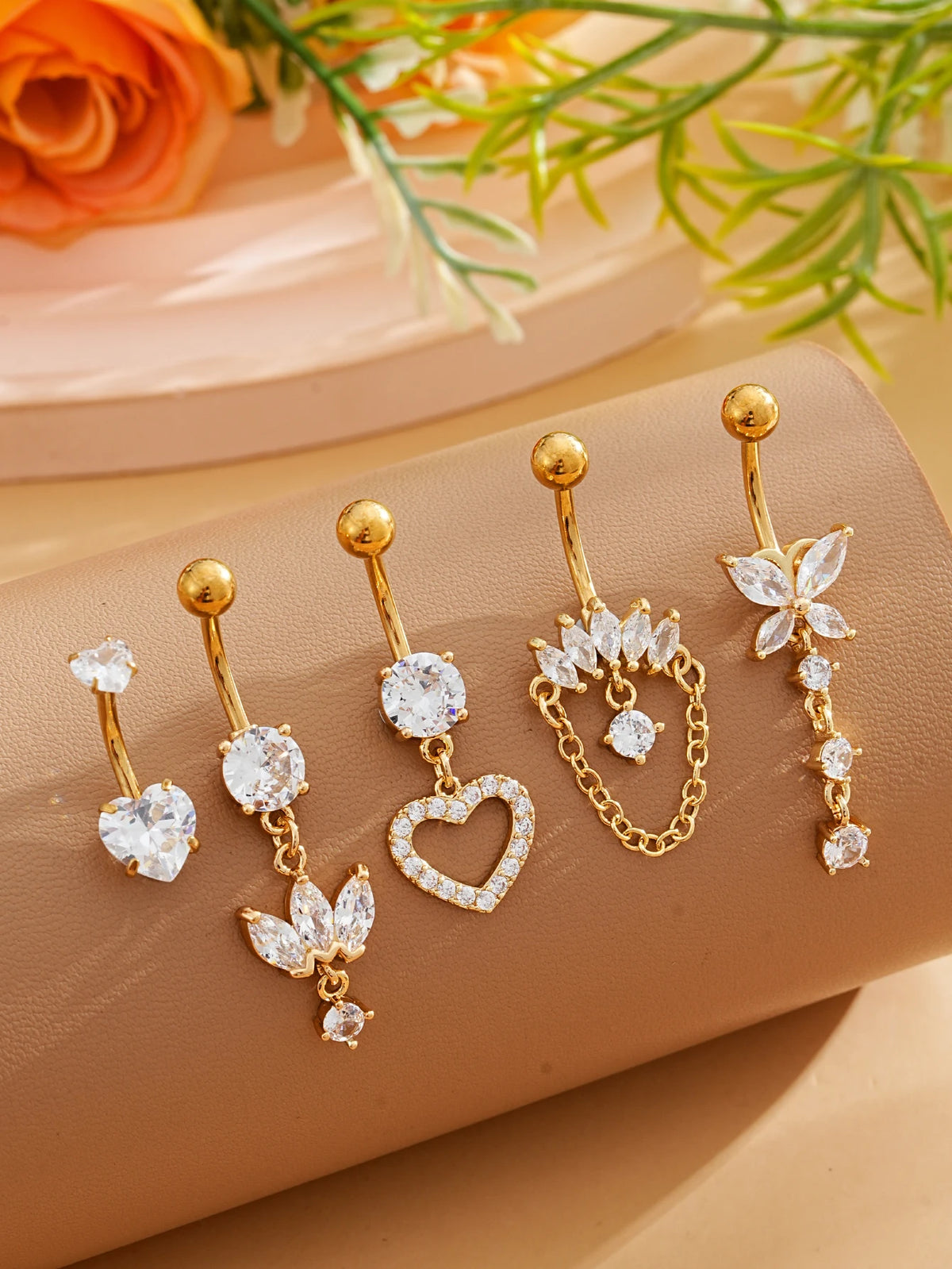 A set of five gold stainless steel dangled belly button rings with diamond accents displayed on a beige surface.
