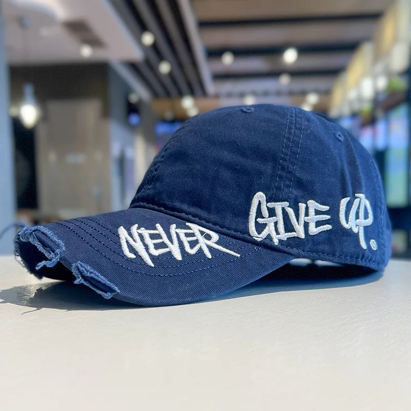 A dark blue vintage washed cotton baseball cap with white embroidered text that reads NEVER GIVE UP.