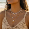 A close-up of a woman wearing a layered gold and pearl necklace with a shell starfish pendant against a beige top.