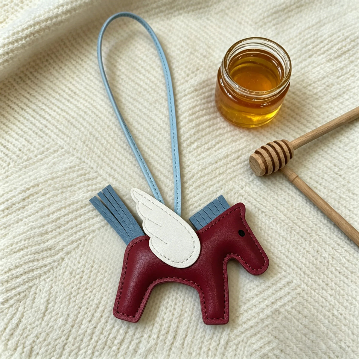 A maroon horse pendant charm with white wings and blue accents rests on a cream-colored fabric next to a small glass jar of h