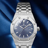 A silver luxury men's watch with a blue dial and luminous markers is displayed against a dark blue fabric background.