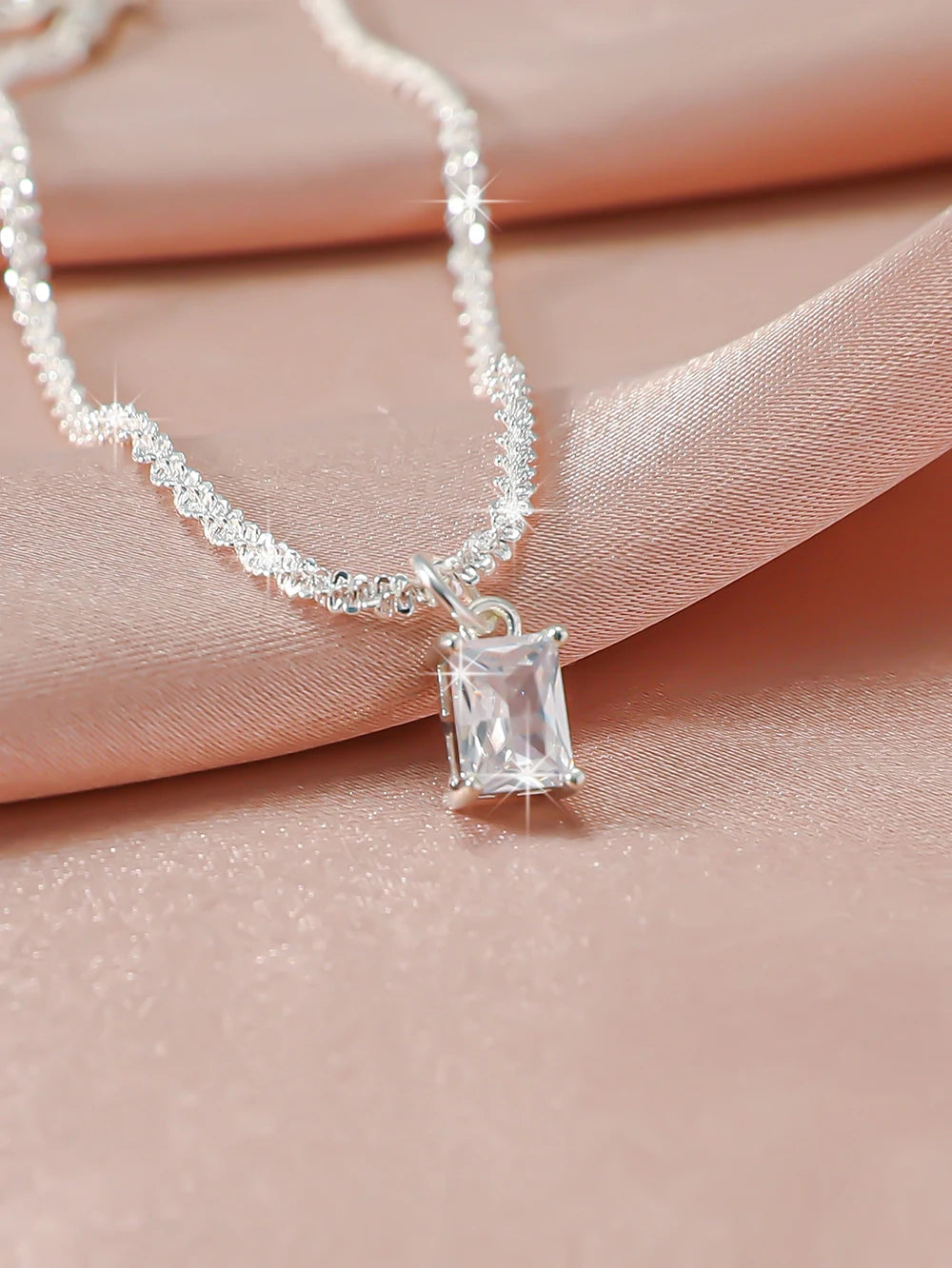 A delicate silver chain necklace with a sparkling zircon heart cross pendant resting on a pink fabric background.