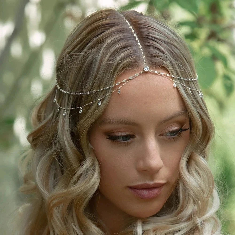 A close-up of a woman with blonde wavy hair wearing a delicate crystal head chain bridal hair accessory.