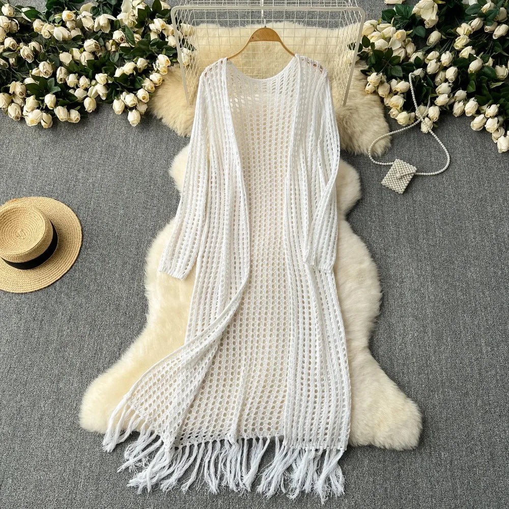 A white knitted cardigan with fringe is displayed on a fluffy beige fur rug surrounded by white flowers.