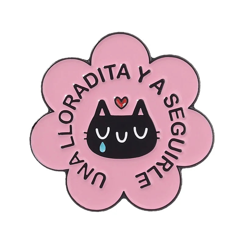 A pink enamel flower brooch featuring the text LLORADITA Y A SEGUIRLE surrounded by a heart and cat design.