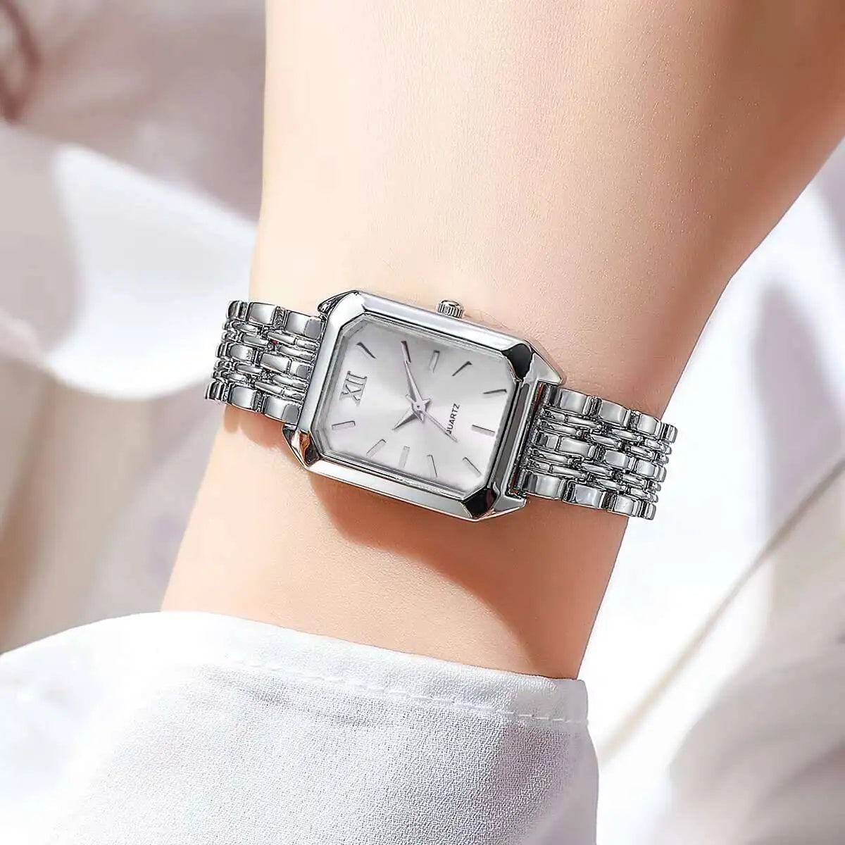 Reloj analógico de cuarzo plateado con esfera cuadrada y pulsera de metal usado en una muñeca.