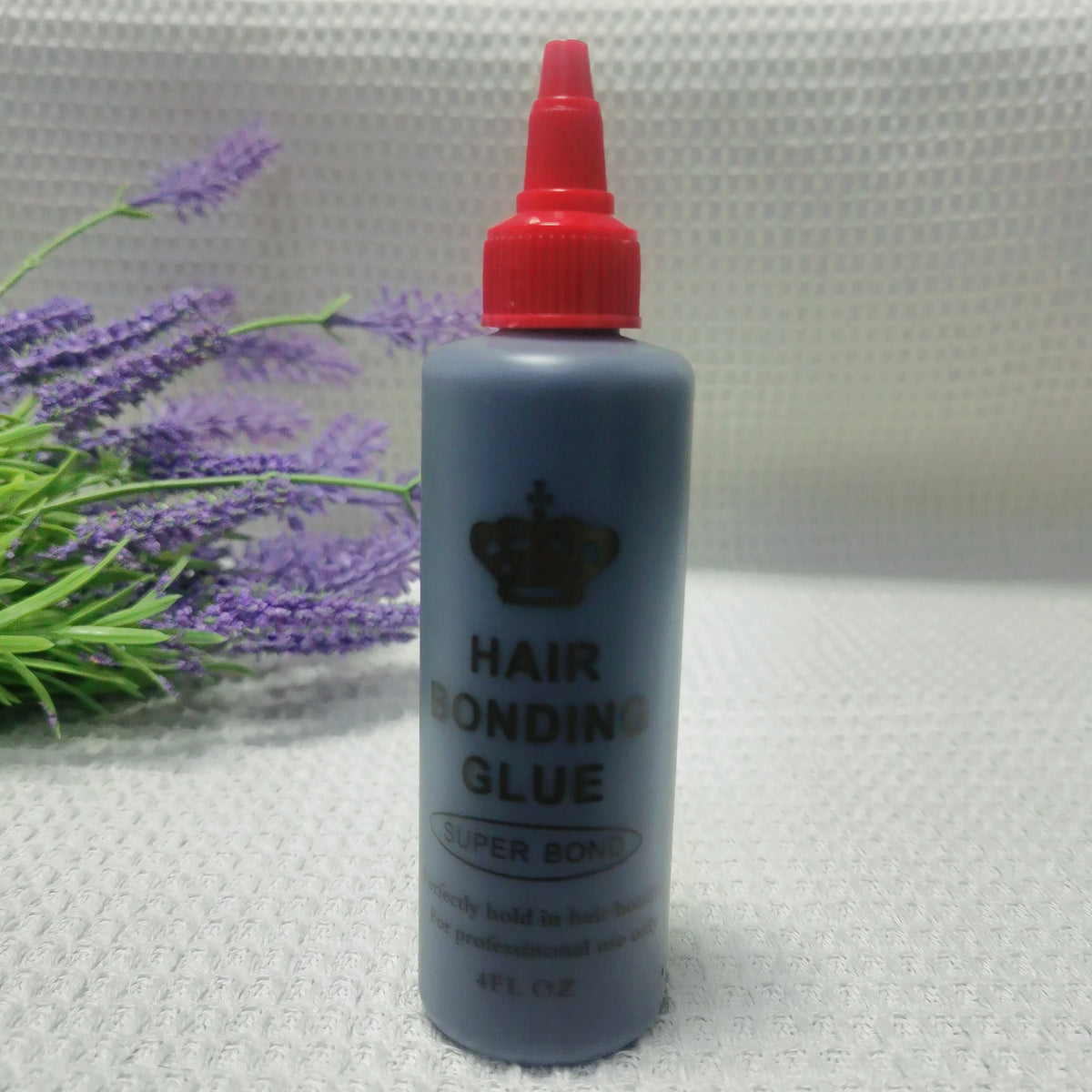 A bottle of professional hair bonding glue with a red cap and dark blue body sits on a light surface next to purple lavender 