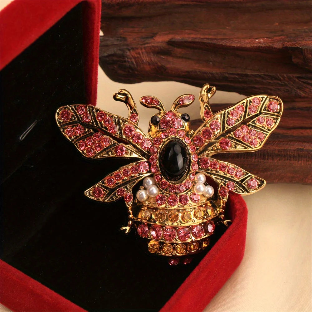 Broche ton or avec motif abeille baroque et insecte, pierres roses et noires dans une boîte de velours rouge.