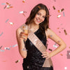 A smiling young woman wearing a sparkly black dress holds an elegant gold sash with the word GRI and a glass of amber liquid 