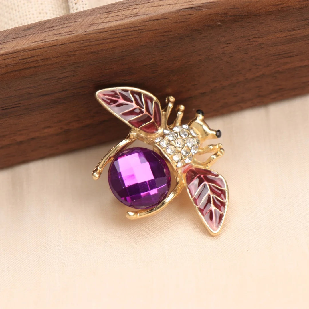 Broche ton or avec motif d'abeille et accents émail rose et violet sur une surface en bois.
