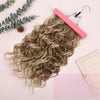 A bundle of light brown curly hair extensions is shown next to a pink hair straightener and a small pinecone on a light pink 