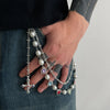 A hand wearing a silver chain with pearl and gemstone crosses is visible against a dark shirt and denim fabric.