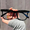 A hand holds a pair of classic black square frames for eye protection against a light gray background.