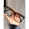 Una mano sostiene unas gafas cuadradas marrón oscuro anti-luz azul contra un fondo gris claro.