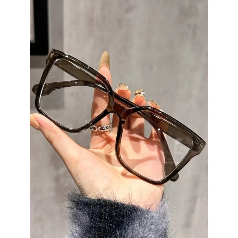 Una mano sostiene unas gafas cuadradas marrón oscuro anti-luz azul contra un fondo gris claro.