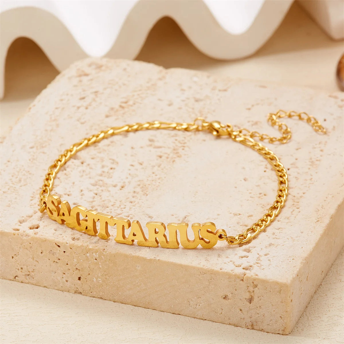 Gold stainless steel zodiac constellation charm bracelet spelling out SAGITARIUS resting on a light colored surface.