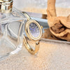 A gold-toned watch with a light blue face and Roman numerals rests on a light beige surface next to a clear glass bottle.