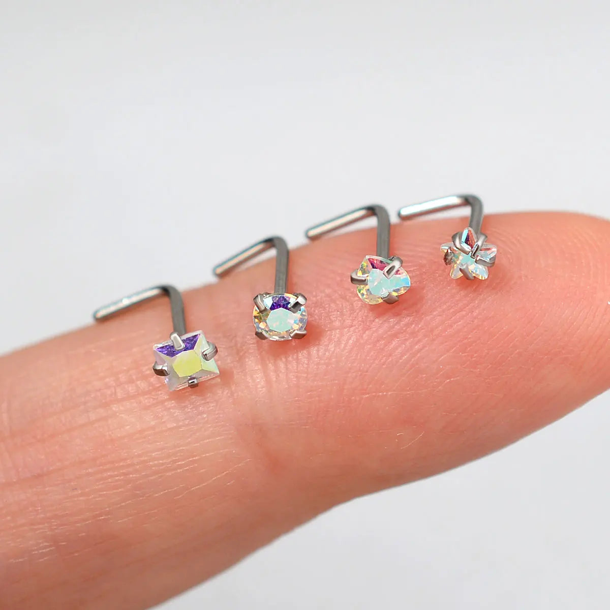 Una mano sostiene varios pendientes en forma de L de acero inoxidable con acentos de gema iridiscente y coloridos.