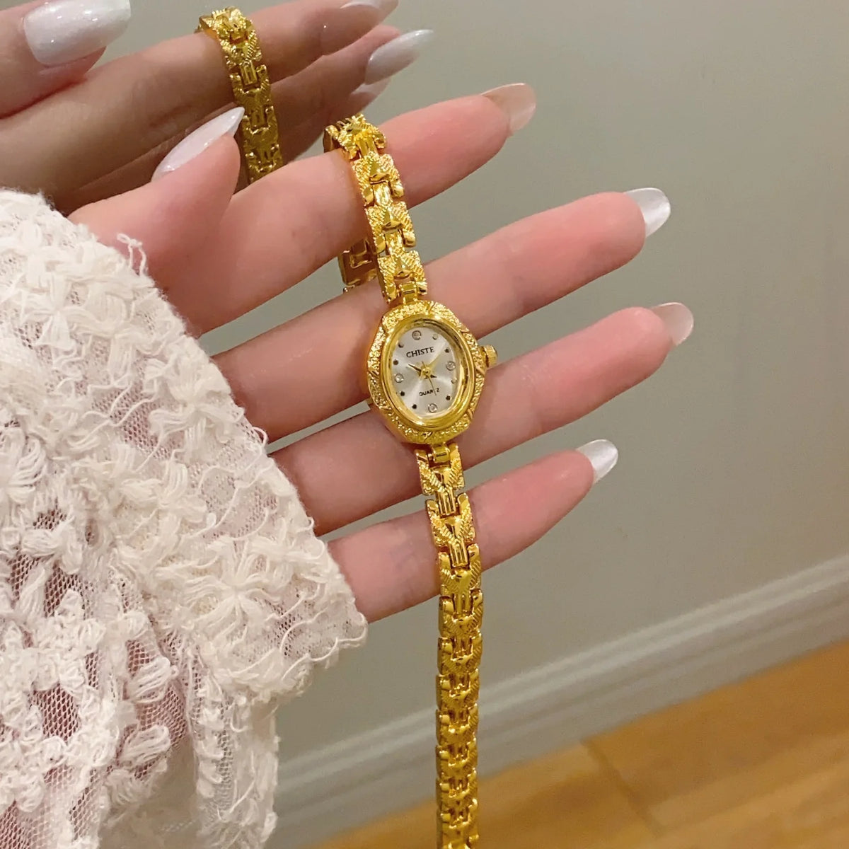 A hand is holding a gold-toned quartz fashion wristwatch with a gold chain and round face against a light background.
