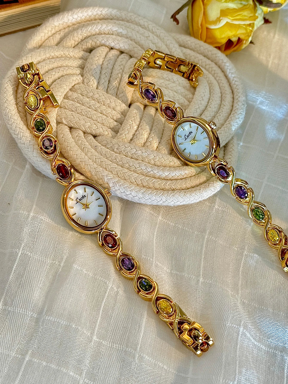 Goldfarbige Modeuhr mit weißem Zifferblatt, verziert mit farbigen Turmalin- und Strassakzenten, liegt auf einem beige...