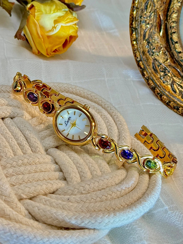 A gold fashion watch adorned with various colored gemstones and rhinestones rests on a beige woven surface.