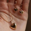 A hand displays a set of dainty gold pearl jewelry including matching heart-shaped earrings and a pendant necklace.