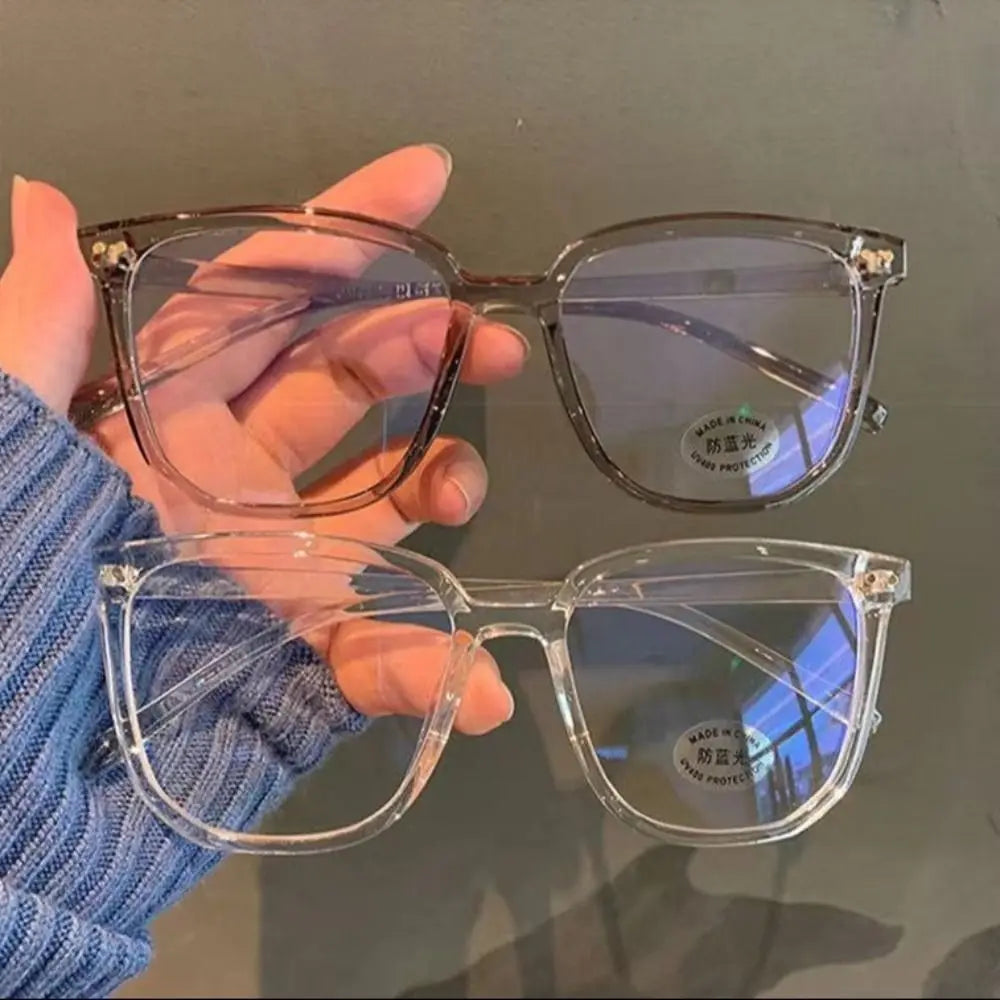 A hand holds a pair of clear, large square anti-blue light spectacles against a dark background.