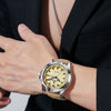 A close-up of a men's quartz watch with a gold-toned case and luminous dial worn on a wrist, paired with a black suit jacket.