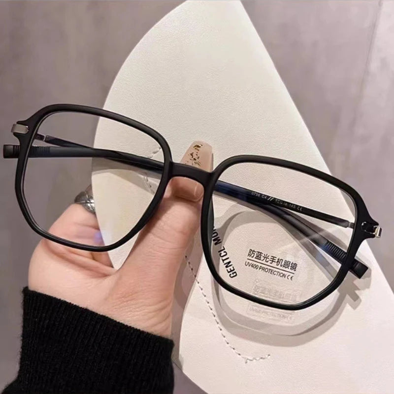 A hand holds a pair of black eyeglasses with dark frames and clear lenses on a white display stand.