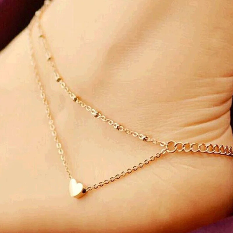 A close-up shot of a delicate gold-toned chain anklet with a heart charm worn on a person's ankle.