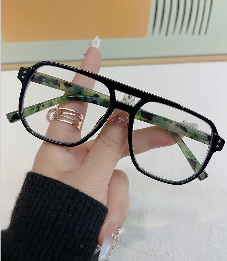 Una mano sostiene unas gafas de lectura con marcos negros y patrón marmolado verde y blanco.