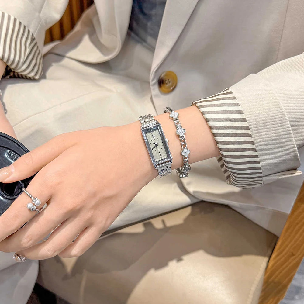 A close-up of a woman's wrist wearing several silver and gold watches and bracelets, next to a light beige blazer.