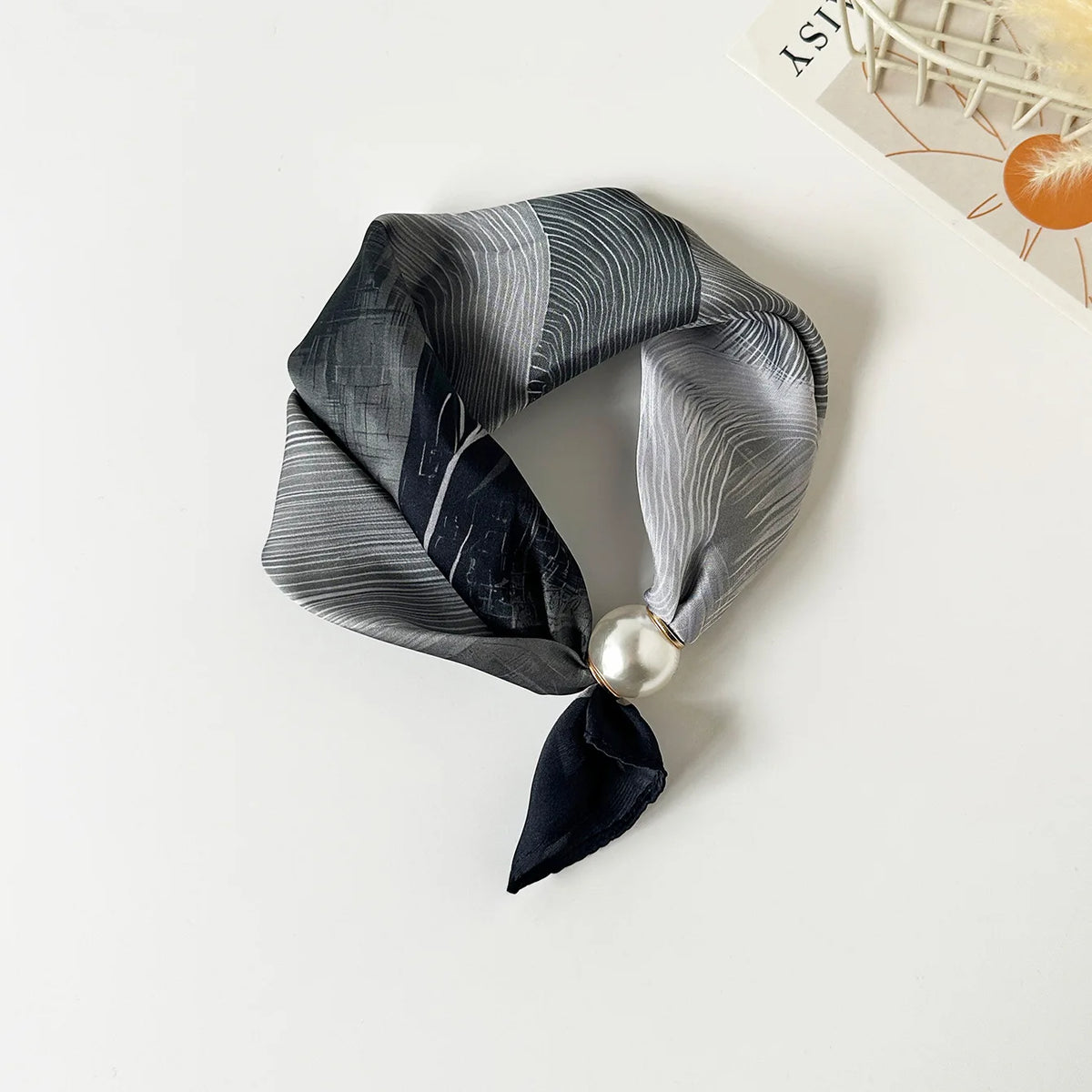 A dark gray silk scarf with a subtle stripe pattern tied in a knot and adorned with a pearl bead.