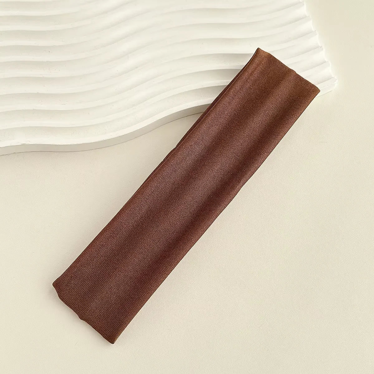 A dark brown fabric headband is laid diagonally on a light surface next to white wavy fabric.
