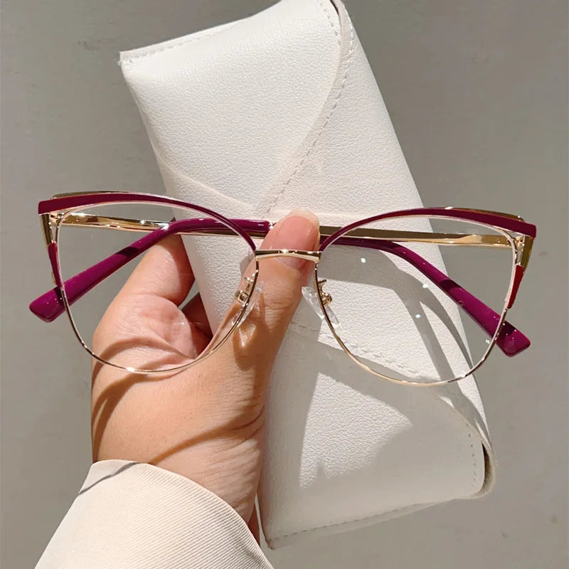 Una mano sostiene unas gafas de computadora con diseño 'cat eye' y marcos morados de metal contra un fondo blanco.