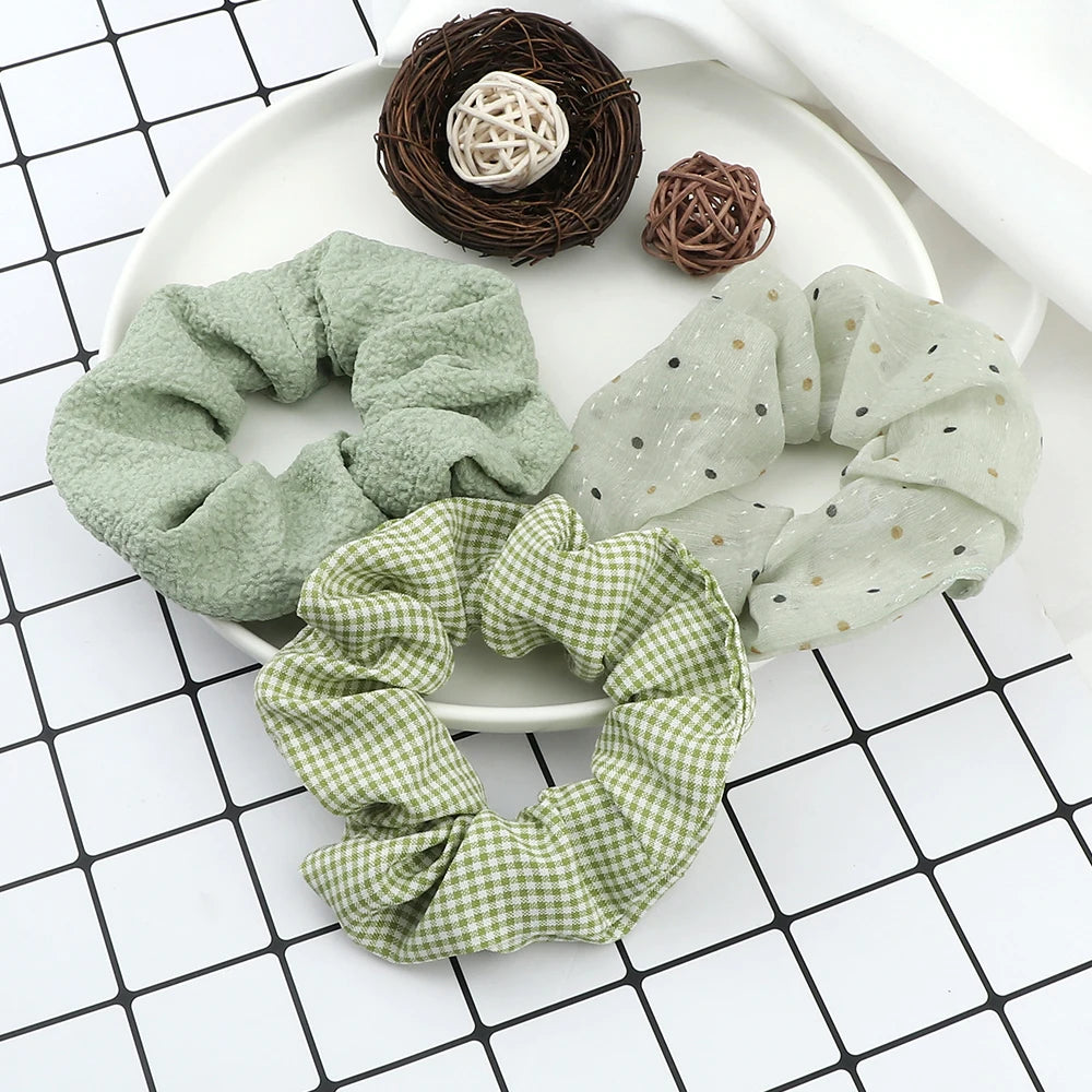 A set of three knitted hair scrunchie headbands in various shades of green and natural brown are displayed on a white plate.