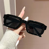 A hand holds a pair of black retro square fashion sunglasses against a light background.