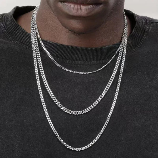 A close-up of a man wearing three layered stainless steel Cuban chain necklaces against a black shirt.