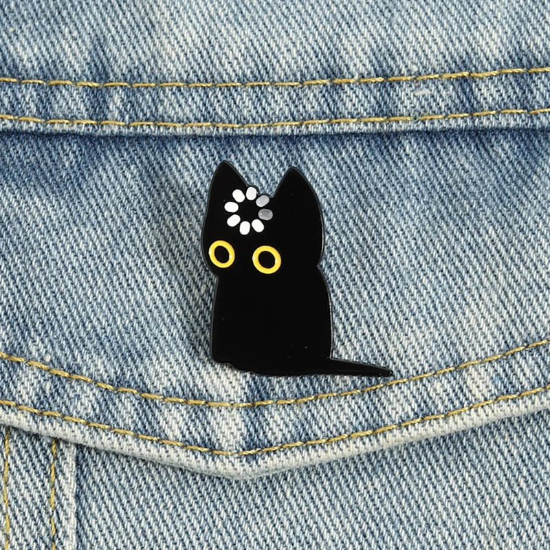 Black cat enamel pin with yellow eyes and white dots pinned on denim fabric.