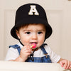A baby wearing a black letter hat with an A and a sun hat is holding a pink lollipop.