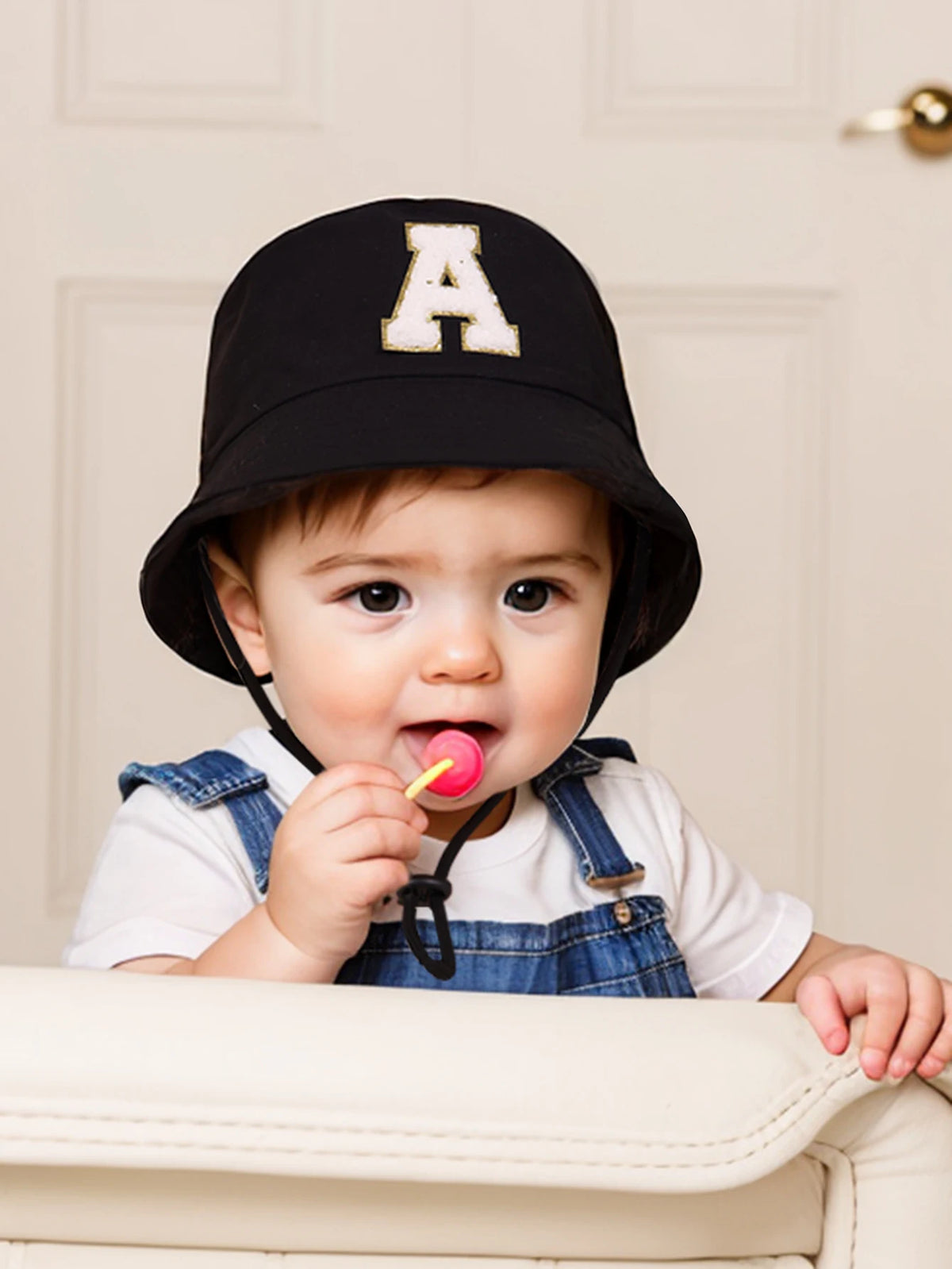 A baby wearing a black letter hat with an A and a sun hat is holding a pink lollipop.
