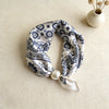 A blue and white silk scarf with a floral print tied in a knot, resting on a beige fabric next to a small vase of dried flowe