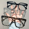 Una mano sostiene una pila de cuatro gafas cuadradas con diferentes monturas y lentes transparentes.