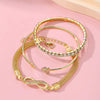 A set of three gold-toned bracelets featuring crystal heart charms displayed on a pink background.
