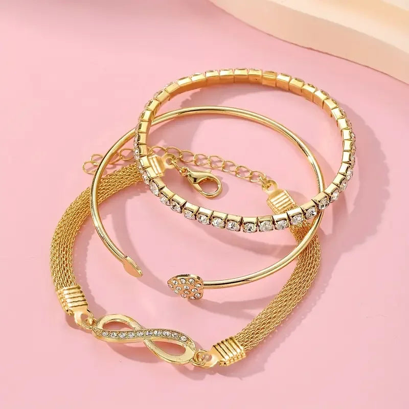 A set of three gold-toned bracelets featuring crystal heart charms displayed on a pink background.