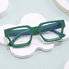 Un par de gafas cuadradas vintage con marco verde oscuro y patillas azules, apoyadas sobre una superficie blanca irregular.