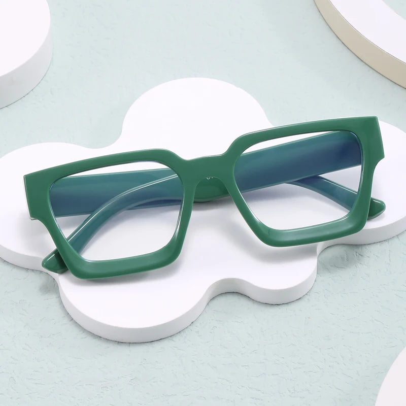 Un par de gafas cuadradas vintage con marco verde oscuro y patillas azules, apoyadas sobre una superficie blanca irregular.