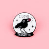 Silueta negra de un pájaro con una joya roja en la cabeza dentro de un pin de esmalte circular con el texto I LOVE y TRINKETS