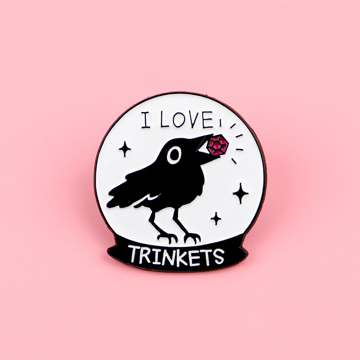 Silueta negra de un pájaro con una joya roja en la cabeza dentro de un pin de esmalte circular con el texto I LOVE y TRINKETS